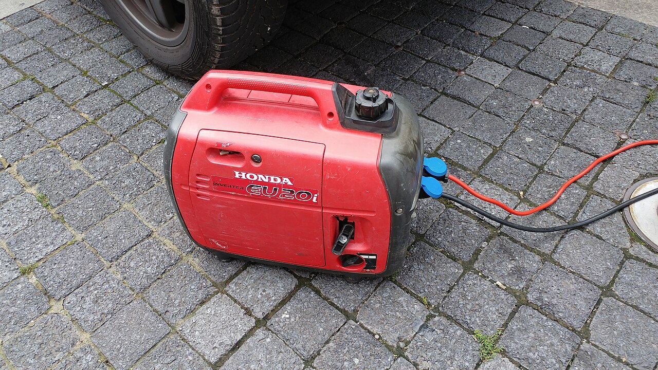 A red Honda EU20i inverter generator on pavers beside a vehicle tire, with cords plugged in.
