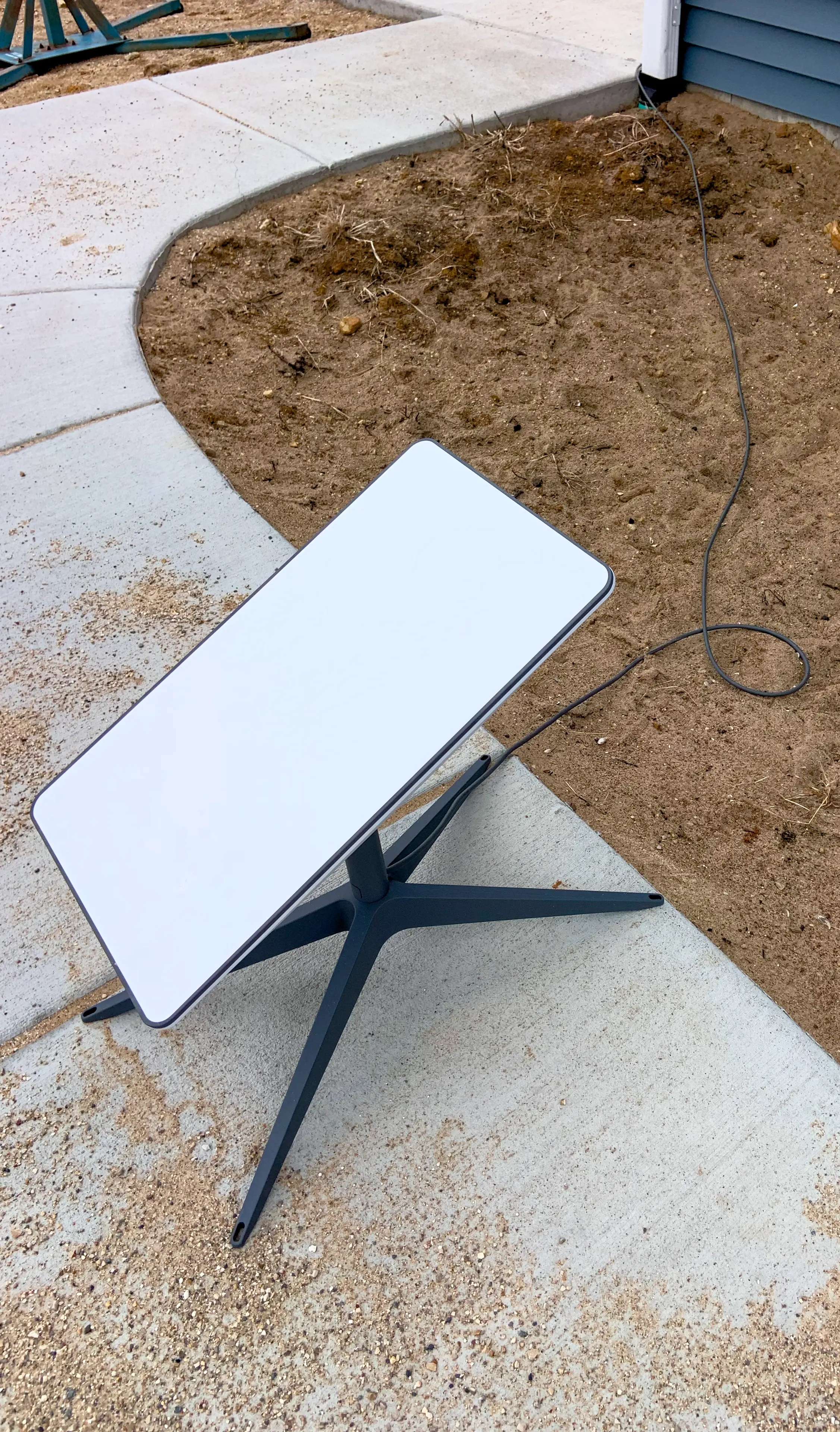 A rectangular Starlink dish on a kickstand, cable coiled on the dirt beside a sidewalk.
